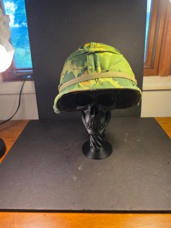 Vietnam Era M1 US Vietnam Era M1 US Military helmet with a reversible "Mitchell" pattern camouflage cover, shown on a display stand.