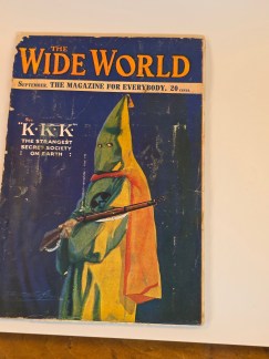 A vintage magazine cover titled "THE WIDE WORLD," showing a Klansman in a green and yellow robe holding a rifle. The text reads, "'K.K.K.' THE STRANGEST SECRET SOCIETY ON EARTH."