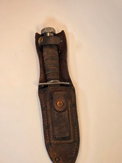 A full view of the vintage Camillus knife in its worn leather sheath, lying on a flat surface.