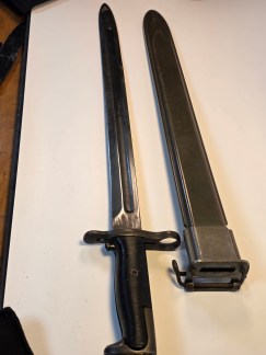 A close-up view of a UFH 1943 dated M1905 bayonet and scabbard. The ricasso markings "O U.S. 1943" and the flaming ordnance bomb are visible.