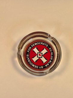A top-down view of a clear glass ashtray with a red, black, and white circular sticker on the bottom. The sticker features the KKK symbol and the words "FOR DEMOCRATS - KU KLUX KLAN - BY DEMOCRATS.