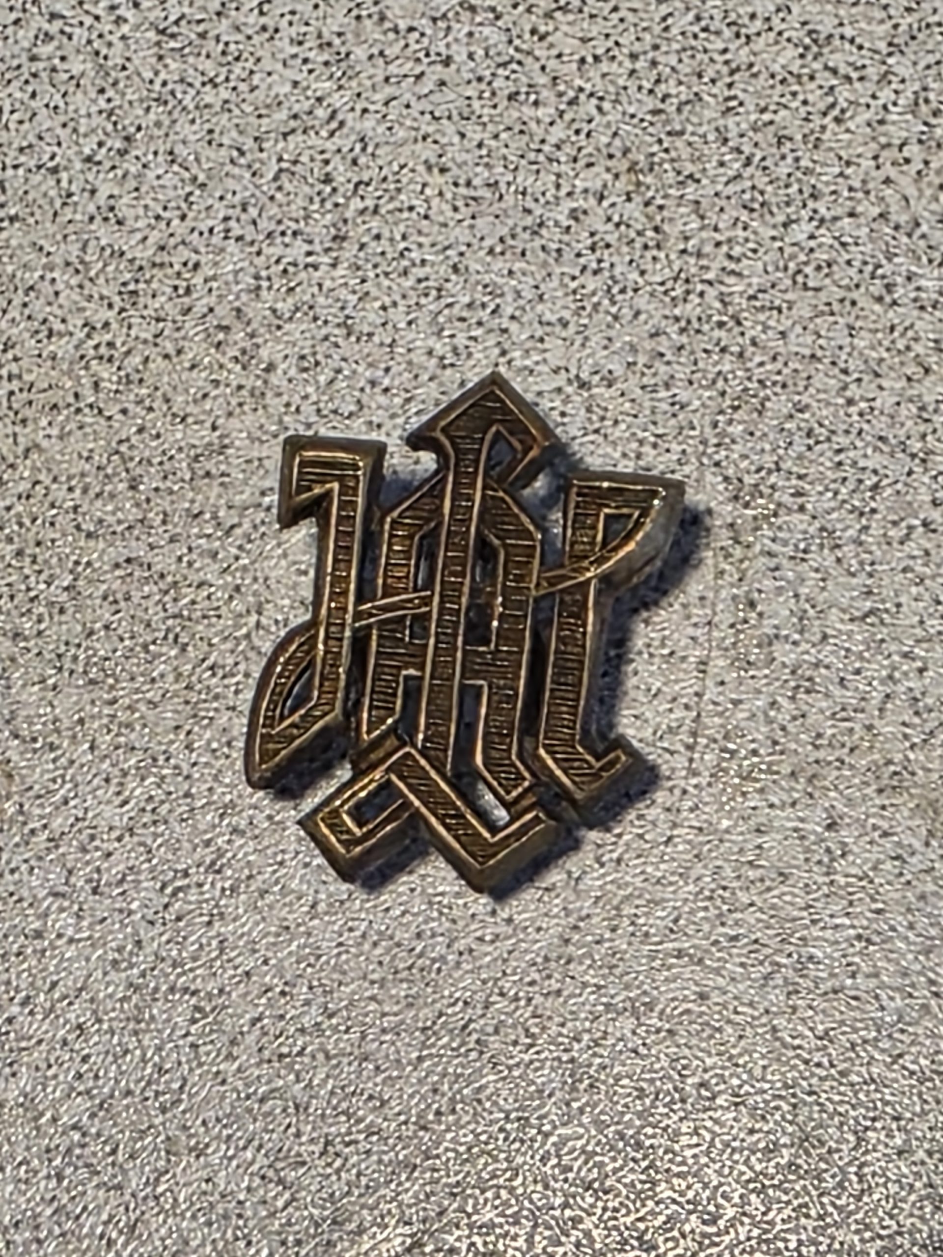 Close-up of a metal WW2 German LSSAH shoulder board cipher with the interlaced letters LAH and mounting prongs on the back.